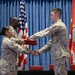 A military ceremony is taking place in an indoor venue where a Marine in desert MARPAT camouflage uniform is receiving a red folder/certificate from a female Marine while shaking hands. A second male Marine stands behind the presenting officer, and a third Marine is seated in the background. The U.S. flag and a Marine Corps unit flag are displayed on either side of the backdrop.