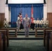 A formal U.S. Marine Corps military ceremony is taking place inside what appears to be a chapel or auditorium on a military installation. A Marine in digital camouflage (MARPAT) uniform stands at attention in the center aisle facing the stage, where additional Marines are positioned near an American flag and a red Marine Corps flag. Several Marines are seated in pews as an audience, and a group of senior personnel are seated on the stage.