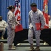 Two U.S. Marines in desert MARPAT camouflage uniforms stand on a stage during what appears to be a formal military ceremony, each holding red folders. They appear to be interacting or exchanging items during the event. Several other Marines are seated in the background, with an American flag and Marine Corps flags visible on stage.