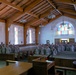 A large group of military personnel in camouflage uniforms are standing at attention inside a military chapel. The chapel features wooden beam ceilings, stained glass windows, hanging lantern-style lights, and wooden pews. The personnel are arranged in rows along both sides of the central aisle, facing a podium at the front.