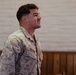 A male service member wearing USMC MARPAT digital camouflage uniform is photographed in profile/three-quarter view, looking upward with mouth open as if speaking or shouting. His name tape reads 'PICERNO' and the branch tape appears to read 'US MARINES'. The setting appears to be an indoor facility with painted brick walls and wood paneling.