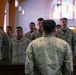 A group of U.S. Marines in woodland/desert MARPAT camouflage uniforms are standing in formation inside a military chapel, appearing to sing or recite something in unison. A single Marine with their back to the camera faces the group, likely in a leadership or conducting role. The setting features stained glass windows and wooden pews, confirming a chapel or religious facility on a military installation.