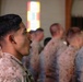 A group of military personnel in desert camouflage uniforms (MARPAT pattern consistent with U.S. Marines) are standing in formation inside what appears to be a chapel or religious facility, indicated by the stained glass window visible in the background. The foreground features a young male service member in profile view, with additional personnel visible in a blurred background. The setting and posture suggest a formal ceremony, memorial service, or religious gathering.