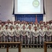 A formal group photograph of U.S. Marines at a Corporal's Course Graduation ceremony. The personnel are arranged in three rows in what appears to be a chapel or auditorium setting, with a projection screen displaying 'Welcome to Corporal's Course Graduation' visible in the background. The group consists of approximately 35-40 service members in desert MARPAT camouflage uniforms.