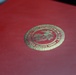 The image shows a red cover or folder embossed with a gold seal of the United States Marine Corps, under the Department of the Navy. The seal features the iconic Eagle, Globe, and Anchor emblem of the USMC rendered in gold foil on a crimson red background. This appears to be an official military document folder or award presentation case.