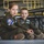 MAINEiacs Return Home Two children are seated on the floor of what appears to be a military aircraft hangar, both wearing olive green flight suits with military patches. The girl on the left has a patch reading '132nd AREFS' and the boy on the right has patches including 'MAINEiacs' and a name tag reading 'ANN ELI MORNEAULT'. Both children are wearing ear protection (earplugs) and appear to be observing something off-camera.