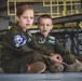 Two children are seated on the floor of what appears to be a military aircraft hangar, both wearing olive green flight suits with military patches. The girl on the left has a patch reading '132nd AREFS' and the boy on the right has patches including 'MAINEiacs' and a name tag reading 'ANN ELI MORNEAULT'. Both children are wearing ear protection (earplugs) and appear to be observing something off-camera.