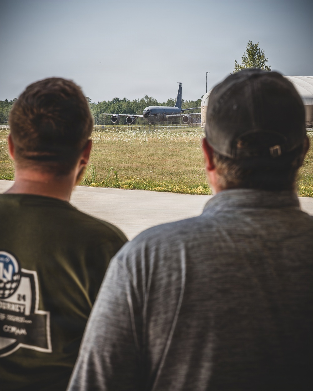 Two individuals are viewed from behind, observing a military aircraft on or near a taxiway at what appears to be a military airfield. The aircraft in the background is a large multi-engine jet, consistent with a tanker or transport type such as a KC-135. The setting includes a grassy infield area, a perimeter fence, and tree line in the background.