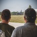 Two individuals are viewed from behind, observing a military aircraft on or near a taxiway at what appears to be a military airfield. The aircraft in the background is a large multi-engine jet, consistent with a tanker or transport type such as a KC-135. The setting includes a grassy infield area, a perimeter fence, and tree line in the background.