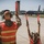 MAINEiacs Return Home Two individuals in military camouflage uniforms and orange safety vests are marshalling a US Air Force aircraft on a tarmac using red signal wands. The larger figure appears to be an adult military personnel while the smaller figure beside them appears to be a child, suggesting a 'bring your child to work' or family day event. A large gray military jet, identifiable as a US Air Force tanker aircraft (likely a KC-135), is taxiing toward them with a control tower visible in the background.