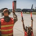 Two individuals in military camouflage uniforms and orange safety vests are marshalling a US Air Force aircraft on a tarmac using red signal wands. The larger figure appears to be an adult military personnel while the smaller figure beside them appears to be a child, suggesting a 'bring your child to work' or family day event. A large gray military jet, identifiable as a US Air Force tanker aircraft (likely a KC-135), is taxiing toward them with a control tower visible in the background.