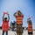 MAINEiacs Return Home A military service member in camouflage uniform and orange safety vest is marshaling a large military aircraft on a flight line, flanked by two children also wearing orange safety vests and holding marshaling wands. All three figures face away from the camera toward the aircraft, with the children mimicking the adult's marshaling signals. This appears to be an educational or community outreach event at a military air base.