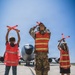 A military service member in camouflage uniform and orange safety vest is marshaling a large military aircraft on a flight line, flanked by two children also wearing orange safety vests and holding marshaling wands. All three figures face away from the camera toward the aircraft, with the children mimicking the adult's marshaling signals. This appears to be an educational or community outreach event at a military air base.