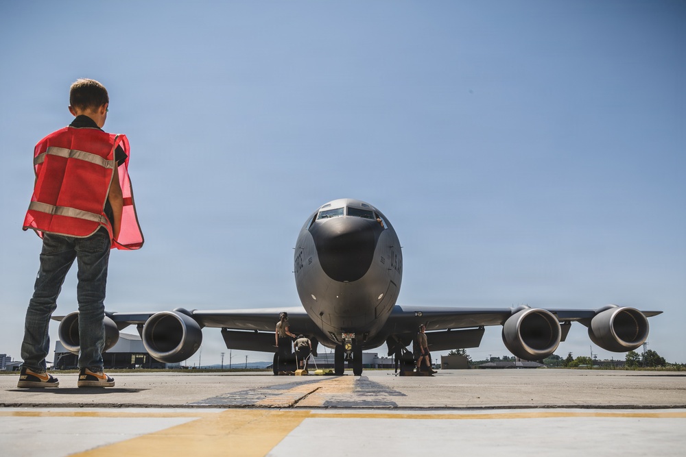 A young boy wearing a red safety vest stands on an airfield tarmac facing a large gray U.S. military aircraft, likely a KC-135 Stratotanker. Ground crew personnel are visible near the nose and landing gear of the aircraft performing pre-flight or maintenance checks. The low-angle shot creates a dramatic perspective emphasizing the scale of the aircraft against a clear blue sky.