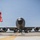 MAINEiacs Return Home A young boy wearing a red safety vest stands on an airfield tarmac facing a large gray U.S. military aircraft, likely a KC-135 Stratotanker. Ground crew personnel are visible near the nose and landing gear of the aircraft performing pre-flight or maintenance checks. The low-angle shot creates a dramatic perspective emphasizing the scale of the aircraft against a clear blue sky.