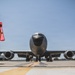 A young boy wearing a red safety vest stands on an airfield tarmac facing a large gray U.S. military aircraft, likely a KC-135 Stratotanker. Ground crew personnel are visible near the nose and landing gear of the aircraft performing pre-flight or maintenance checks. The low-angle shot creates a dramatic perspective emphasizing the scale of the aircraft against a clear blue sky.