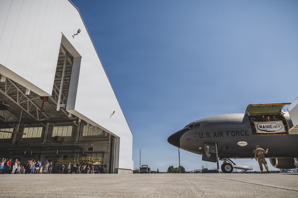 A U.S. Air Force aircraft, marked with 'MAINEiacs' nose art and serial number 3513, is positioned on a tarmac outside a large military hangar. A crowd of civilians is gathered near the hangar entrance, suggesting a public event such as an open house or homecoming ceremony. A uniformed military personnel member stands near the aircraft's nose gear, appearing to direct or greet attendees.