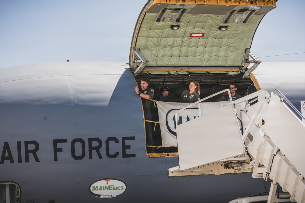 Several military personnel in flight suits are visible inside the open cargo door of a U.S. Air Force aircraft, likely a C-17 Globemaster or similar large transport plane. One male crew member is waving while a female crew member is smiling broadly, suggesting an arrival or departure event. A 'MAINEiacs' sticker with a University of Tennessee logo is visible on the fuselage.