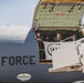 Several military personnel in flight suits are visible inside the open cargo door of a U.S. Air Force aircraft, likely a C-17 Globemaster or similar large transport plane. One male crew member is waving while a female crew member is smiling broadly, suggesting an arrival or departure event. A 'MAINEiacs' sticker with a University of Tennessee logo is visible on the fuselage.