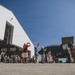 A military homecoming or deployment ceremony is taking place on an airfield tarmac, with civilians and family members gathered to greet or send off military personnel. Soldiers in camouflage uniforms are descending the stairs of a large U.S. Air Force aircraft, while crowds of civilians photograph and embrace them. The event is occurring on a sunny day beside a large aircraft hangar.