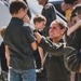 A female military aviator in an olive green flight suit kneels down to greet a young boy, smiling emotionally in what appears to be a homecoming reunion. Red, white, and blue flowers are visible on the ground beside her, and other military personnel and children are visible in the background near what appears to be an aircraft stairway. The scene strongly suggests a military deployment return ceremony.