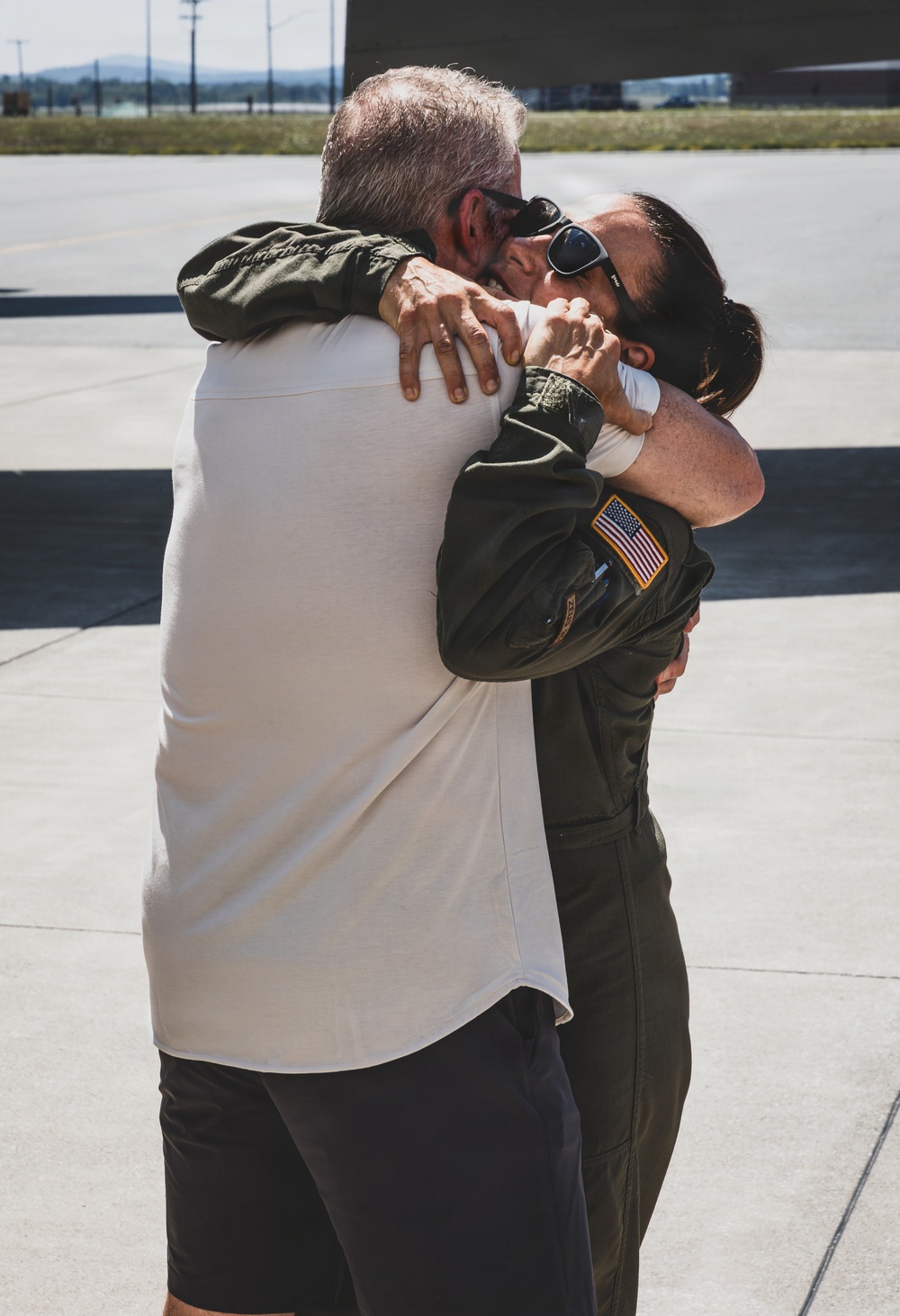 Two individuals are sharing an emotional embrace on what appears to be a military airfield tarmac. One person is wearing a dark olive green flight suit with a U.S. flag patch on the sleeve, suggesting they are military aviation personnel. The setting and attire indicate this may be a homecoming or departure reunion moment.