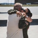 Two individuals are sharing an emotional embrace on what appears to be a military airfield tarmac. One person is wearing a dark olive green flight suit with a U.S. flag patch on the sleeve, suggesting they are military aviation personnel. The setting and attire indicate this may be a homecoming or departure reunion moment.