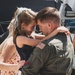 A U.S. military servicemember in an olive green flight suit is holding a young girl, their foreheads touching in an emotional reunion moment. A woman in a floral dress stands beside them, her hand resting on the child. The scene appears to be a military homecoming event, likely at an airfield or naval facility.
