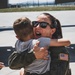 A person wearing an olive green military flight suit with a U.S. flag patch and a name tape reading 'ZEUS 95' is joyfully embracing a young child on what appears to be a military airfield tarmac. The individual is expressing intense emotion with an open-mouthed smile, suggesting a reunion after a deployment or extended absence. The setting and attire are consistent with a U.S. military homecoming event.
