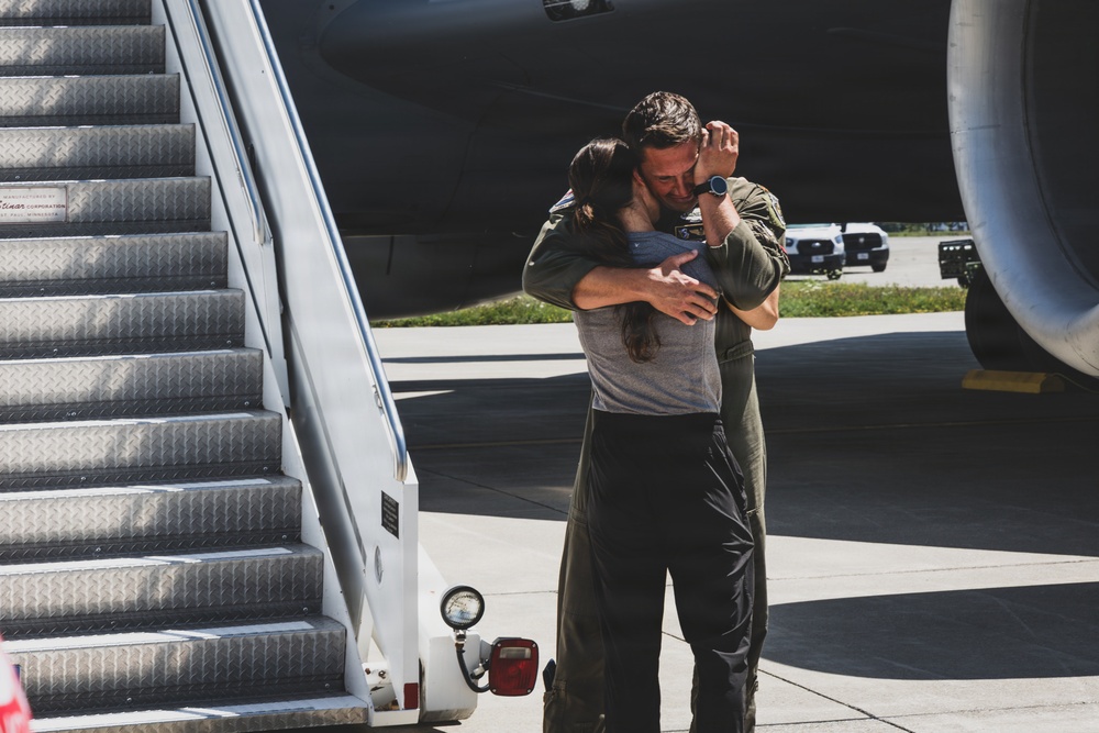Two individuals are embracing near the base of an aircraft boarding stairway on a tarmac. One person is wearing an olive green flight suit, suggesting a military or aviation context. The emotional embrace appears to be a reunion or farewell scene at an airfield.