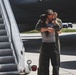 Two individuals are embracing near the base of an aircraft boarding stairway on a tarmac. One person is wearing an olive green flight suit, suggesting a military or aviation context. The emotional embrace appears to be a reunion or farewell scene at an airfield.
