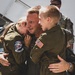 A military airman in an olive green flight suit embraces two children, also dressed in matching flight suits, in what appears to be a homecoming or reunion on an airfield tarmac. The adult wears a US flag patch and a unit patch reading '132nd AREFS' is visible on one of the children's suits. The emotional scene captures a joyful family reunion in a military aviation setting.
