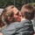 MAINEiacs Return Home A close-up photograph showing an adult with blonde hair in a ponytail kissing a young child on the cheek while embracing them. The child is wearing a gray quilted short-sleeve shirt. The setting appears to be outdoors with green trees visible in the background.