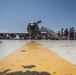 A large military jet aircraft, likely a KC-135 Stratotanker or similar tanker/transport aircraft, is parked on a tarmac with a boarding stairway attached. A group of civilians and military personnel are gathered near the aircraft, suggesting a public open house, air show, or family day event. The photo is taken from a low ground-level angle with a yellow taxiway line leading toward the aircraft.