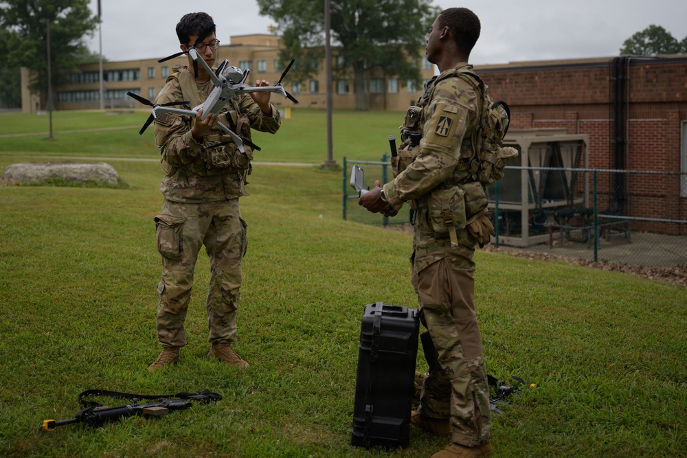 Two soldiers in U.S. Army OCP (Operational Camouflage Pattern) uniforms are preparing or inspecting a commercial quadcopter drone on a grassy area of what appears to be a military installation. One soldier holds the drone while the other holds what appears to be a remote controller. Equipment including a hard-shell carrying case and a rifle are visible on the ground nearby.