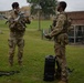 Two soldiers in U.S. Army OCP (Operational Camouflage Pattern) uniforms are preparing or inspecting a commercial quadcopter drone on a grassy area of what appears to be a military installation. One soldier holds the drone while the other holds what appears to be a remote controller. Equipment including a hard-shell carrying case and a rifle are visible on the ground nearby.