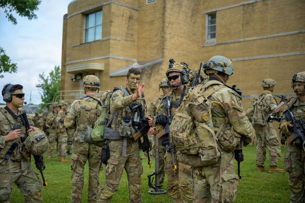 A group of military personnel in full combat gear are assembled on a grass field in front of a tan brick building. The soldiers are wearing Operational Camouflage Pattern (OCP) uniforms and are equipped with rifles, tactical vests, helmets, and communication headsets. One soldier appears to be briefing or gesturing to others, suggesting a pre-mission or post-mission assembly.