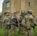 A group of military personnel in full combat gear are assembled on a grass field in front of a tan brick building. The soldiers are wearing Operational Camouflage Pattern (OCP) uniforms and are equipped with rifles, tactical vests, helmets, and communication headsets. One soldier appears to be briefing or gesturing to others, suggesting a pre-mission or post-mission assembly.