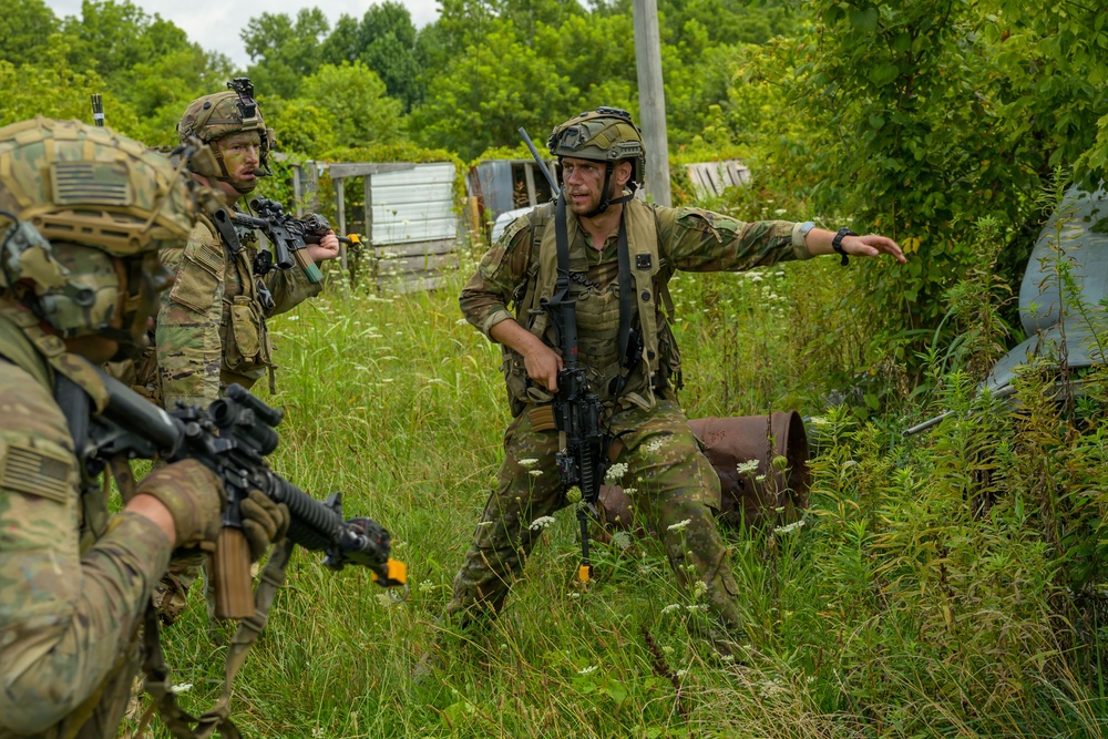 Three soldiers in camouflage uniforms are conducting what appears to be a military training exercise in an overgrown field area. One soldier in the center is gesturing directionally while holding a rifle, appearing to give tactical hand signals or directions to teammates. The setting includes vegetation, abandoned structures, and rusty barrels, consistent with a simulated urban or rural combat training environment.