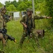 Three soldiers in camouflage uniforms are conducting what appears to be a military training exercise in an overgrown field area. One soldier in the center is gesturing directionally while holding a rifle, appearing to give tactical hand signals or directions to teammates. The setting includes vegetation, abandoned structures, and rusty barrels, consistent with a simulated urban or rural combat training environment.