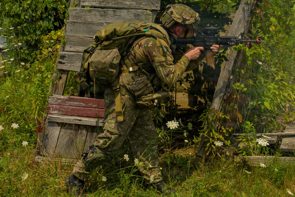 A soldier in full combat gear is moving tactically through overgrown vegetation near a dilapidated wooden structure. The individual is aiming a rifle while advancing in a crouched position, suggesting a training exercise or operational movement. The setting appears to be a rural or abandoned area during daylight hours.