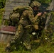 A soldier in full combat gear is moving tactically through overgrown vegetation near a dilapidated wooden structure. The individual is aiming a rifle while advancing in a crouched position, suggesting a training exercise or operational movement. The setting appears to be a rural or abandoned area during daylight hours.
