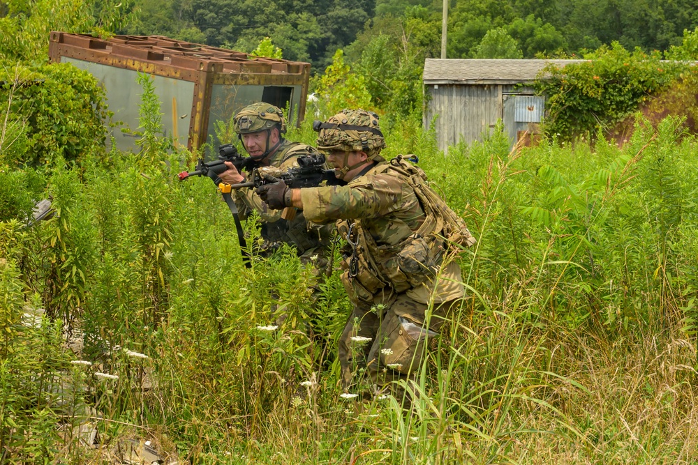 Two soldiers in camouflage uniforms and helmets are moving through dense vegetation in what appears to be a military training exercise. They are carrying rifles with visible training/blank-firing attachments (orange and yellow muzzle devices), indicating this is a simulated combat drill rather than live combat. The setting includes overgrown terrain with abandoned structures in the background.