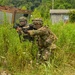 Two soldiers in camouflage uniforms and helmets are moving through dense vegetation in what appears to be a military training exercise. They are carrying rifles with visible training/blank-firing attachments (orange and yellow muzzle devices), indicating this is a simulated combat drill rather than live combat. The setting includes overgrown terrain with abandoned structures in the background.