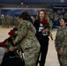 A military ceremony taking place in an indoor gymnasium or arena, where soldiers in U.S. Army OCP (Operational Camouflage Pattern) uniforms are presenting bouquets of red roses to civilians. The event appears to be a formal recognition or farewell ceremony, with rows of uniformed soldiers seated in the background. A civilian woman in a black outfit is receiving roses while another soldier stands nearby smiling.