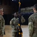 A U.S. Army soldier identified by nametape as 'COAXUM' holds a furled dark blue guidon or unit colors with gold fringe during what appears to be a military change of command or activation ceremony. The ceremony is taking place on a hardwood gymnasium or arena floor, with other uniformed soldiers standing nearby. The soldier wears OCP (Operational Camouflage Pattern) uniform with a unit patch featuring a starburst design on the left shoulder.