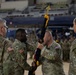 A U.S. Army change of command or activation ceremony is taking place inside what appears to be an indoor arena or gymnasium. Two soldiers in the center are exchanging a unit guidon (flag on a staff with gold fringe and finial), which is the traditional symbolic act of transferring command authority. Additional soldiers stand on either side as witnesses to the formal military ceremony.