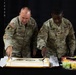 Two U.S. Army soldiers in OCP (Operational Camouflage Pattern) uniforms are cutting a ceremonial cake at what appears to be a military celebration or anniversary event. The soldier on the left is identified by name tape as 'McKENNON' and holds a senior NCO rank, while the soldier on the right is identified as 'COAXUM' and wears a Colonel's rank insignia. A fruit platter is visible to the right of the cake.