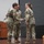 USARIEM COC/Relinquishment of Responsibility 2026 Three U.S. Army soldiers in OCP (Operational Camouflage Pattern) uniforms are on a stage during what appears to be a formal military ceremony. Two soldiers in the foreground are shaking hands, suggesting a change of command, promotion, or award presentation. Military flags and a podium are visible in the background.