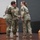 USARIEM COC/Relinquishment of Responsibility 2026 Three U.S. Army soldiers in OCP (Operational Camouflage Pattern) uniforms are on a stage during what appears to be a military ceremony, likely a change of command or award presentation. Two soldiers are shaking hands while a third stands closely behind, suggesting a formal handoff or recognition event. Military flags and a podium are visible in the background.